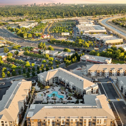 Sacramento living at it's finest Aerial view of a modern apartment complex in Natomaswith a beautifully landscaped courtyard and pool, surrounded by lush greenery and vibrant cityscape at sunrise.